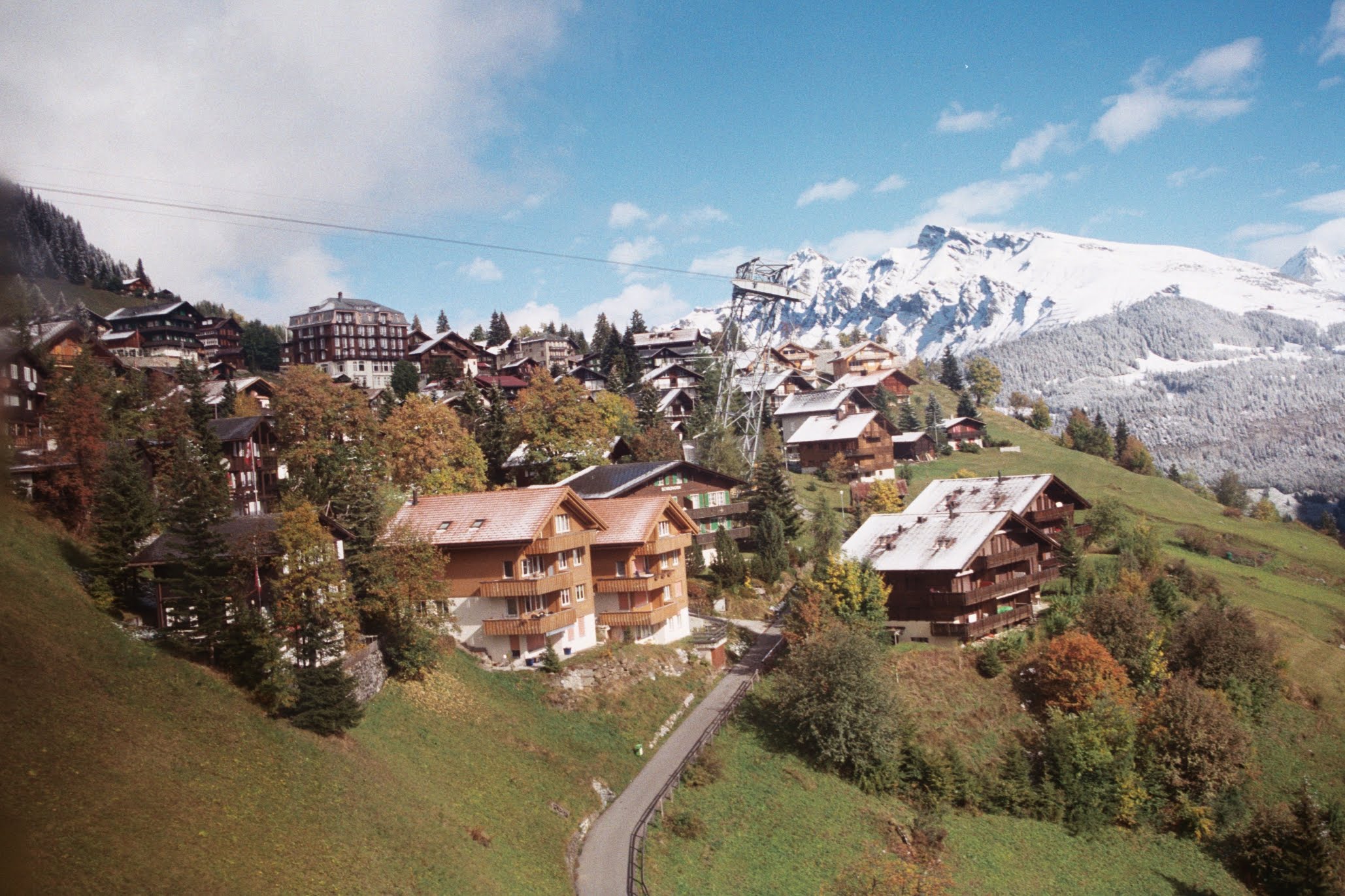 A scenic mountain village with wooden chalets scattered along a green hillside, dusted with early snow. In the background, tall snow-covered peaks rise under a blue sky with light clouds, creating a stunning contrast of autumn and winter in the Swiss Alps.