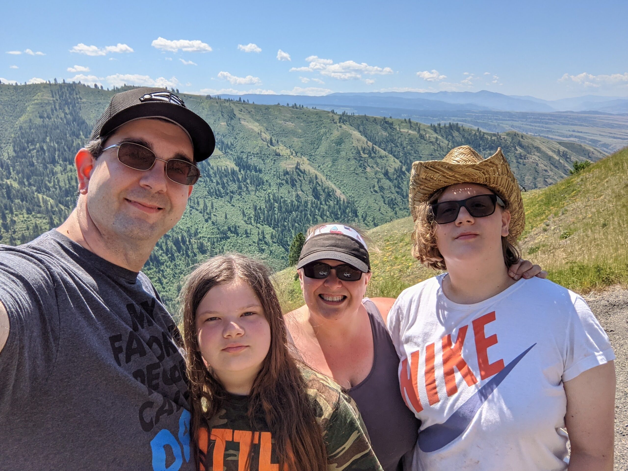 A smiling family of four takes a selfie on a sunny day with a stunning mountain valley in the background. The parents and two children wear casual summer clothes, sunglasses, and hats, standing close together on a hillside with green ridges and distant blue mountains behind them.